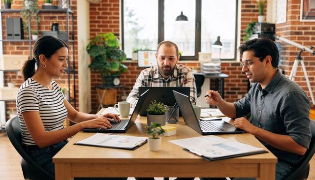 Three coworkers collaborating and reviewing charts on laptops in modern brick office