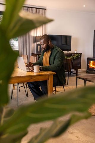 African american man working from home at dining table with laptop and cozy fireplace