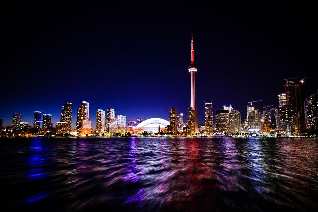 Toronto skyline lit up at night with vibrant lights reflecting on the water. Iconic CN Tower visible among the towering buildings with a dark blue sky. Ideal for travel guides, promotional content related to Toronto, or editorial use highlighting Canada's largest city.