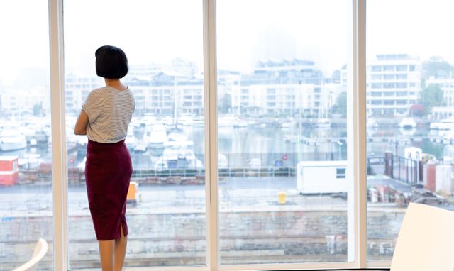 Businessperson standing with arms crossed, looking out of office window at marina and cityscape. Ideal for use in business, corporate, and professional contexts, emphasizing contemplation, strategy, and modern work environments.