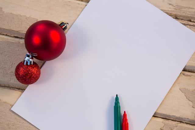 Christmas baubles and blank paper with red and green markers on a wooden plank. Perfect for holiday greeting card designs, festive invitations, or Christmas craft projects. Ideal for use in seasonal advertising, social media posts, or blog articles about holiday preparations and decorations.