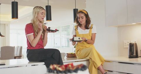 Two friends enjoying cake while sitting on kitchen island in bright modern kitchen