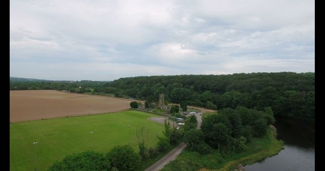 An aerial view captures a serene landscape with a historic ruin beside a river, surrounded by lush trees and open fields. The image evokes a sense of tranquility and the enduring beauty of nature alongside historical architecture.