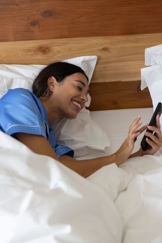 Young woman relaxing in bed using smartphone with wooden headboard and white bedding