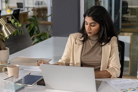Indian businesswoman working at laptop reviewing printed charts in modern office