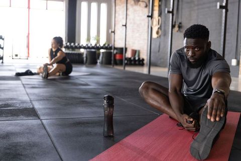 Black man stretching on red mat in industrial gym with blurred partner and kettlebells