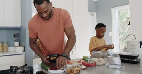 African american father and son preparing healthy meals and packing containers on kitchen island