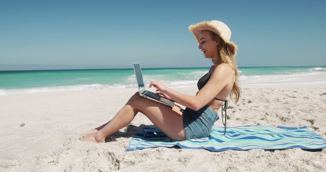 Young woman sitting on beach towel, working on laptop near ocean on bright sunny day. Perfect for depicting themes of remote work, freelancing, digital nomad lifestyle, travel, summer vacations, and work-life balance. Could be used for advertisements, website headers, blog posts about productivity, work while traveling, and summer promotions.