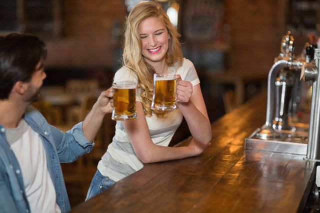Young woman and male friend toasting beer mugs at a pub. Both are smiling and enjoying their time together. Ideal for use in advertisements for bars, pubs, or social events, as well as articles about nightlife, friendship, and leisure activities.