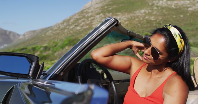 Woman wearing sunglasses driving convertible in sunny landscape. Ideal for concepts of travel, summer, freedom, and adventure. Useful for marketing campaigns involving cars, road trips, and outdoor activities.