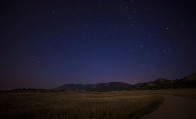 Starry night sky full of stars over a peaceful countryside meadow with mountains in the distance. Suitable for use in projects related to nature, outdoor activities, astronomy, or peaceful and serene settings.