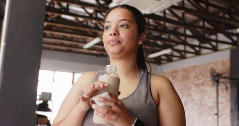 Young woman pausing during workout holding glass bottle in loft gym with smartwatch