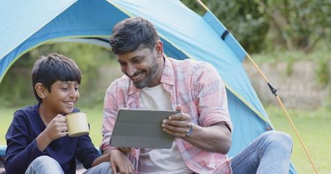 Father and son in joyful moment experiencing outdoor camping while using a tablet. The laughter and comfortable atmosphere highlight family bonding and modern technology amid nature, indicating an engaging outdoor adventure. Ideal for content related to parenting, family leisure activities, outdoor promotions, technology integration in family life, as well as camping tips and advertisements.