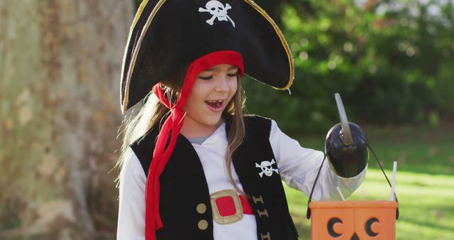 Child enjoying Halloween outdoors in park while holding candy bucket wearing pirate costume. Perfect for use in Halloween promotions, kids' costume ideas, holiday celebrations, or festive events marketing.