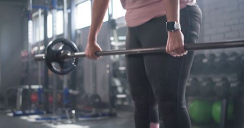Woman deadlifting loaded barbell wearing smartwatch and pink top in gym strength training
