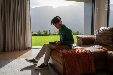 Asian man using tablet with headphones on leather chaise in sunlit living room