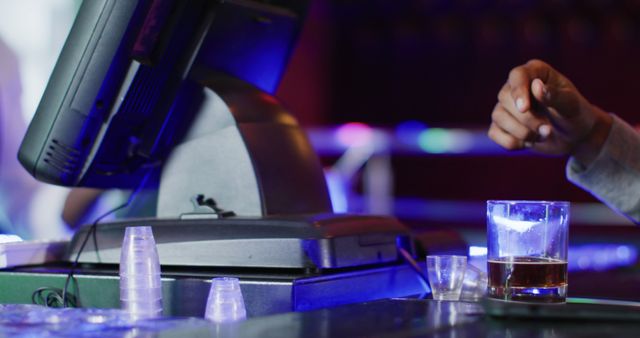 Close-up view of customer drinking at a bar counter. Hand seen next to touchscreen terminal, with a glass of liquor nearby. Ideal for concepts of nightlife, relaxation, bar ambiance, and social activities.