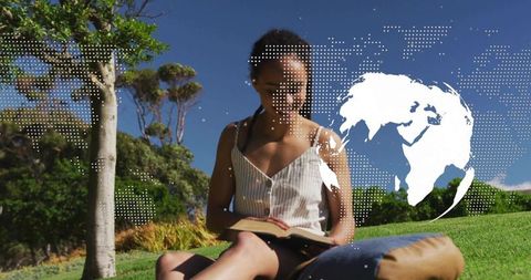 Young woman reading paperback on sunny park lawn with dotted world map overlay