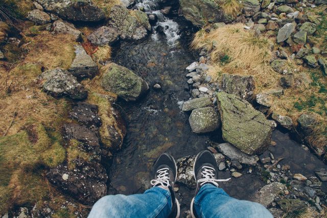 Feet in casual hiking shoes and jeans dangling above a scenic rocky creek. Captures a sense of adventure and exploration. Suitable for travel blogs, outdoor activity promotions, hiking gear advertisements, and nature photography collections.