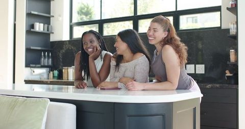 Three diverse friends leaning on kitchen island laughing together in bright modern home