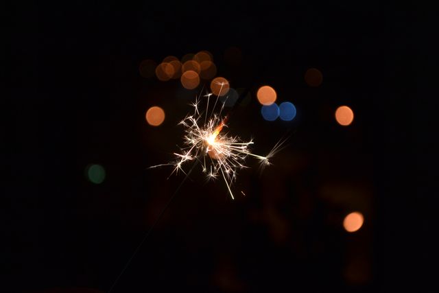 Hand holding a lit sparkler in the dark with glowing bokeh lights in the background. Ideal for themes on celebrations, parties, festivals, New Year's Eve, or any festive occasion. Great for advertisements, blogs, social media posts, and event promotions highlighting fun and excitement.