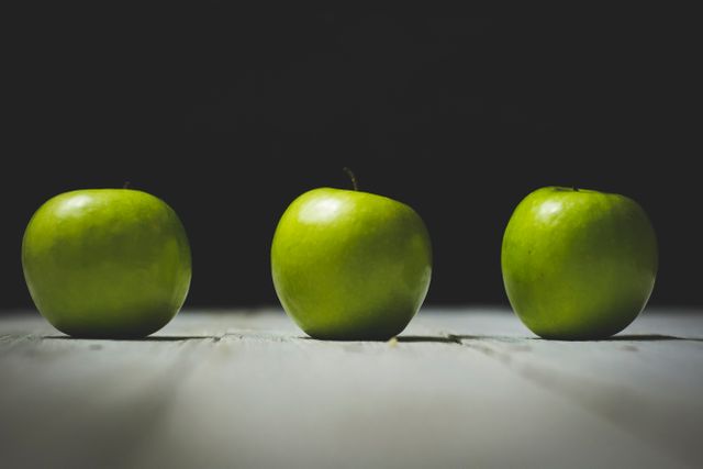 This photo showcases three green apples placed on a wooden table against a dark background, evoking simplicity and minimalism. Suitable for use in articles and advertisements focused on healthy eating, diet, organic produce, or minimalist lifestyle concepts.