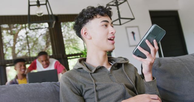 Teenage boy using a voice assistant on a smartphone while sitting on a couch. Two friends in the background are working on a laptop and laughing together. Suitable for themes of technology use among youth, casual home life, and communication. Also ideal for illustrating modern family or friend interaction and diverse communities.