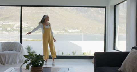 Mature woman standing at panoramic window wearing yellow overalls with waterfront view