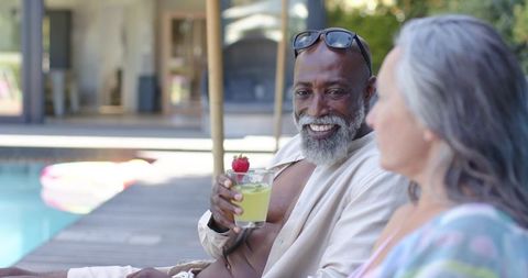 Smiling diverse senior couple relaxing poolside sipping cocktails at luxury resort