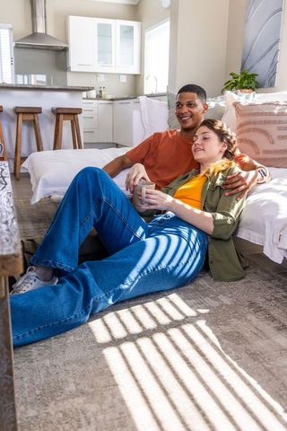 Multiracial couple lounging on living room floor enjoying coffee in sunlit open-plan home