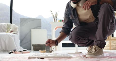 Indian artist crouching in sunlit loft applying palette knife paint on canvas with mountain view