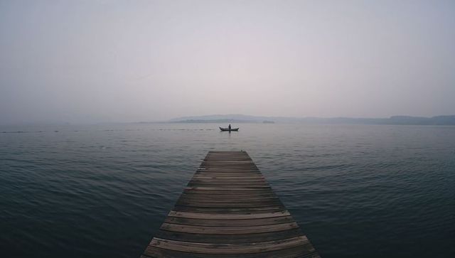 Wooden pier leading into misty lake toward solitary rowboat over calm horizon