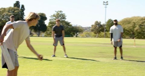 Youth team practicing cricket on sunlit grassy park pitch, batting and fielding action