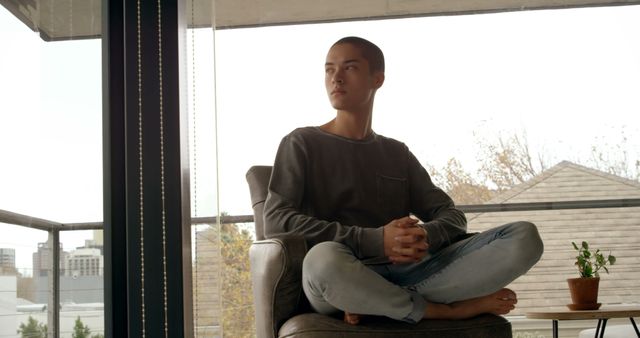 Young man sitting in a comfortable chair at home in an urban setting, engaging in meditation. Suitable for themes on relaxation, mindfulness, mental health, urban living, or promoting a peaceful lifestyle.