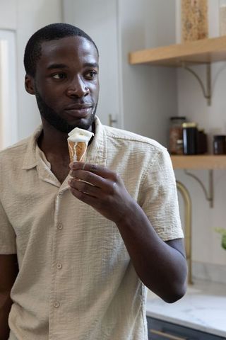 African american man enjoying melting vanilla ice cream cone in modern kitchen lifestyle