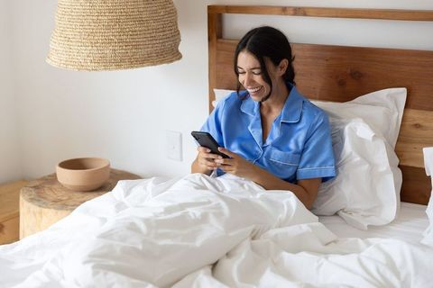 Woman smiling and texting in bed wearing blue satin pajamas in modern rustic bedroom