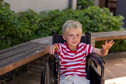 Blond boy sitting in wheelchair reaching out in sunny courtyard, accessibility, inclusion