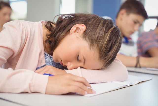 Young girl resting her head on a desk in a classroom, appearing tired and asleep. Ideal for illustrating concepts related to student fatigue, academic stress, and the importance of sleep for children. Useful for educational articles, school-related content, and discussions on student well-being.