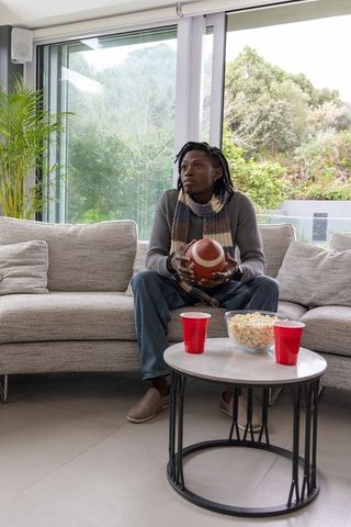 African american man holding football, sitting on modern sectional, watching game at home