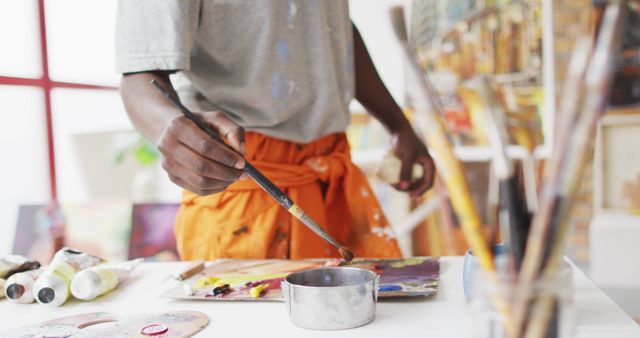 A close-up of an artist's hand holding a paintbrush while working on a painting in a studio. Art supplies such as palettes, paint tubes, and brushes are visible around, adding to the creative atmosphere. This image is ideal for use in art-related materials, promotional content for art studios or workshops, websites or blogs about painting and creativity.