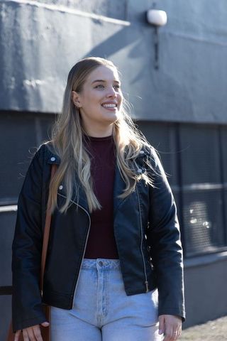 Young woman smiling on sunny urban sidewalk wearing leather jacket and high waist jeans