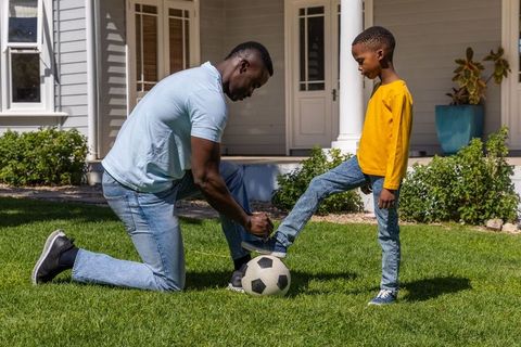 African american father tying son's soccer shoe on green lawn with soccer ball