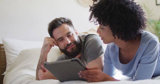 Image of happy diverse couple relaxing at home, lying on bed using tablet. Happiness, communication, inclusivity, free time, togetherness and domestic life.