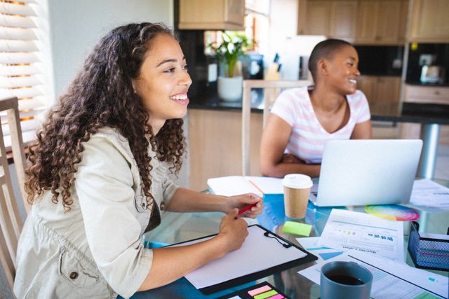 Young biracial businesswoman sitting with female colleague at dining table inside home. unaltered, people, friends, and business concept.