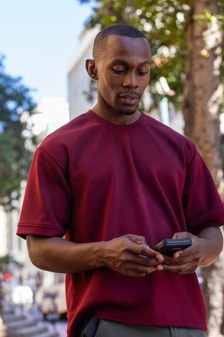 African american man checking smartphone on sunlit city sidewalk wearing burgundy tee