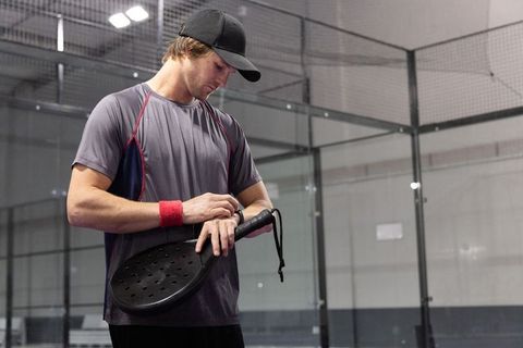 Padel player checking smartwatch while holding paddle inside glass court