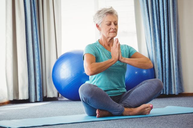 Senior woman practicing meditation in lotus position on yoga mat at home. Blue exercise ball in background. Ideal for promoting senior fitness, mindfulness, home exercise routines, and healthy lifestyle for elderly.
