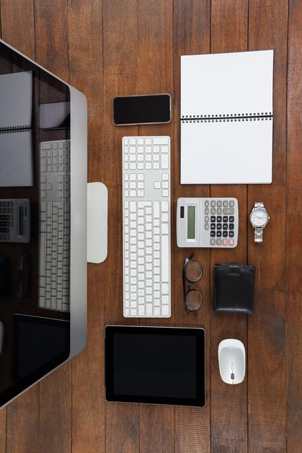 Overhead view of a well-organized office desk featuring a computer monitor, keyboard, tablet, smartphone, notebook, calculator, glasses, wristwatch, wallet, and a mouse. Useful for illustrating workplace organization, modern office setup, tech-savvy environment, productivity, and business tasks.