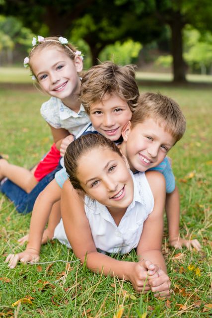 Group of happy children stacking on each other while lying on grass in a park on a sunny day. Ideal for use in advertisements, educational materials, and websites promoting outdoor activities, family fun, and childhood joy.