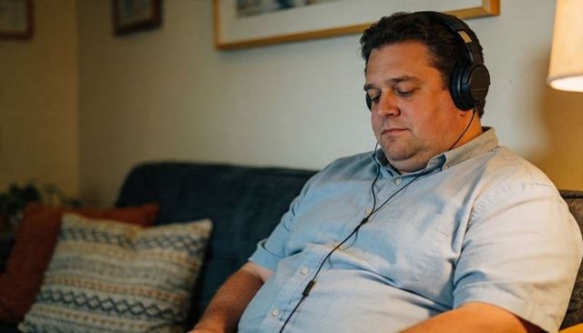 Middle-aged man relaxing on sofa listening to music with wired headphones under warm lamp